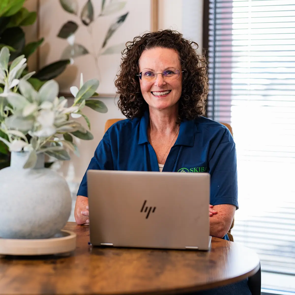 A woman with curly hair and glasses sits at a wooden table, smiling at the camera while using an HP laptop. She is wearing a blue polo shirt, managing small business bookkeeping Stillwater OK. A potted plant and a window are in the background. -Skiba Bookkeeping