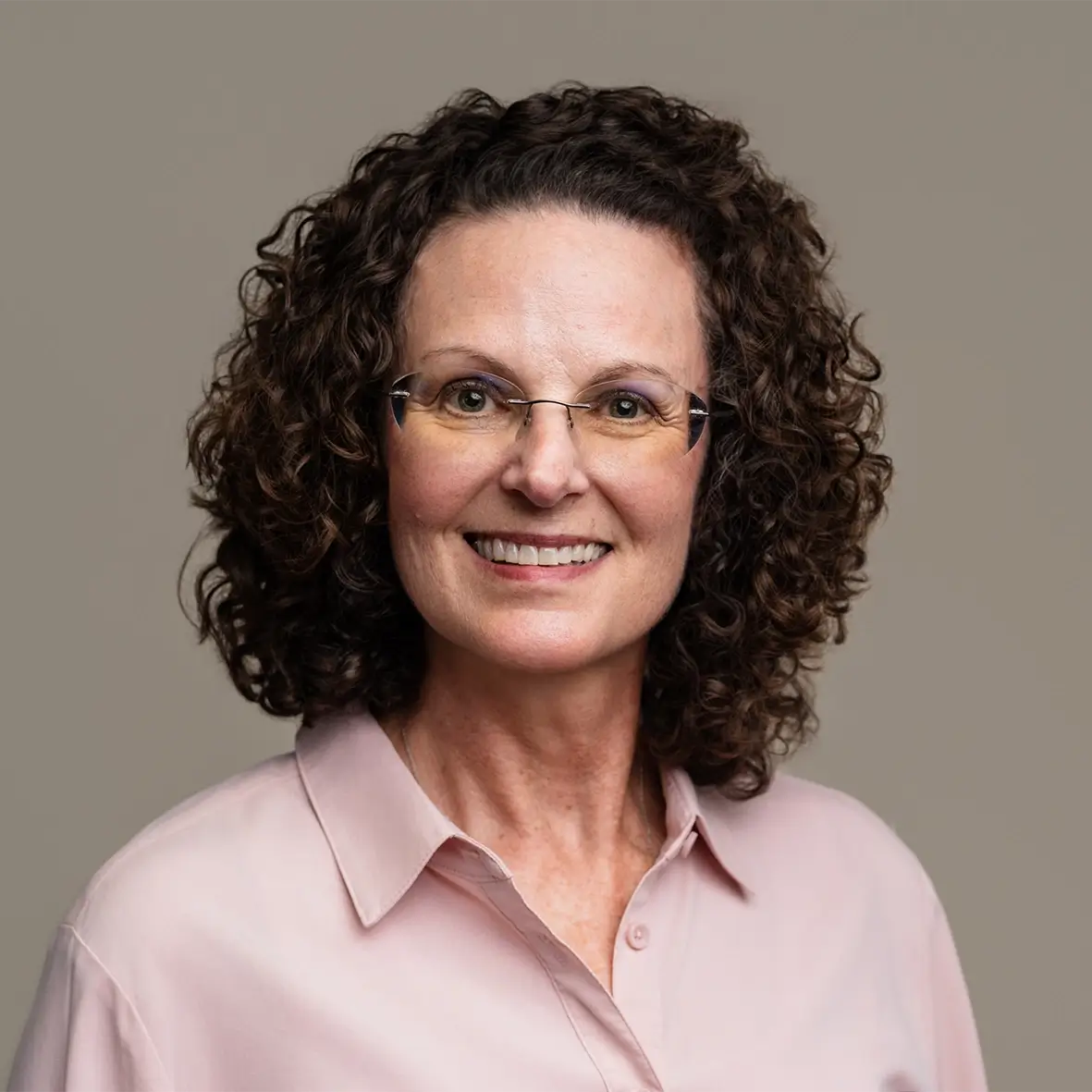 A woman with short, curly brown hair and glasses smiles at the camera. She is wearing a light pink collared shirt—ready to assist with small business bookkeeping Stillwater OK—standing in front of a plain, beige background. -Skiba Bookkeeping