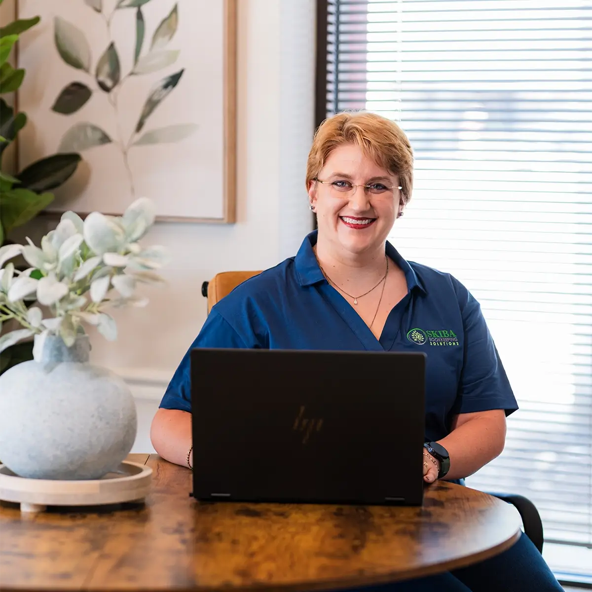 A bookkeeper with short blond hair and glasses sits at a round wooden table, working on a laptop. She wears a blue collared shirt with an embroidered logo and smiles at the camera. A vase with greenery sits on the table nearby. -Skiba Bookkeeping