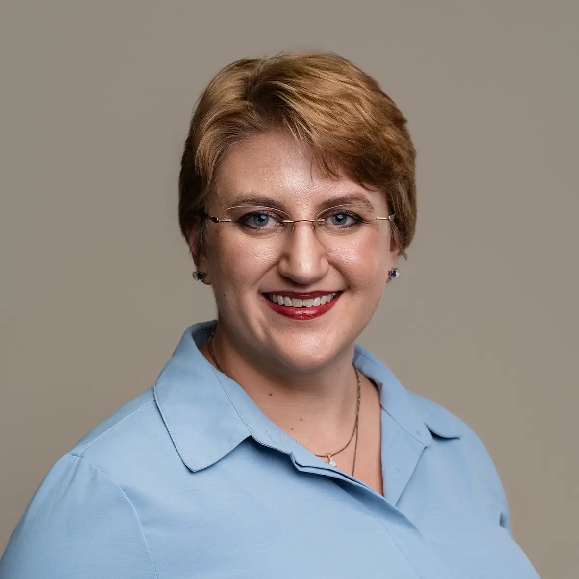 A person with short light brown hair, wearing glasses and a light blue collared shirt, is smiling at the camera against a plain beige background, representing small business bookkeeping Stillwater OK. -Skiba Bookkeeping