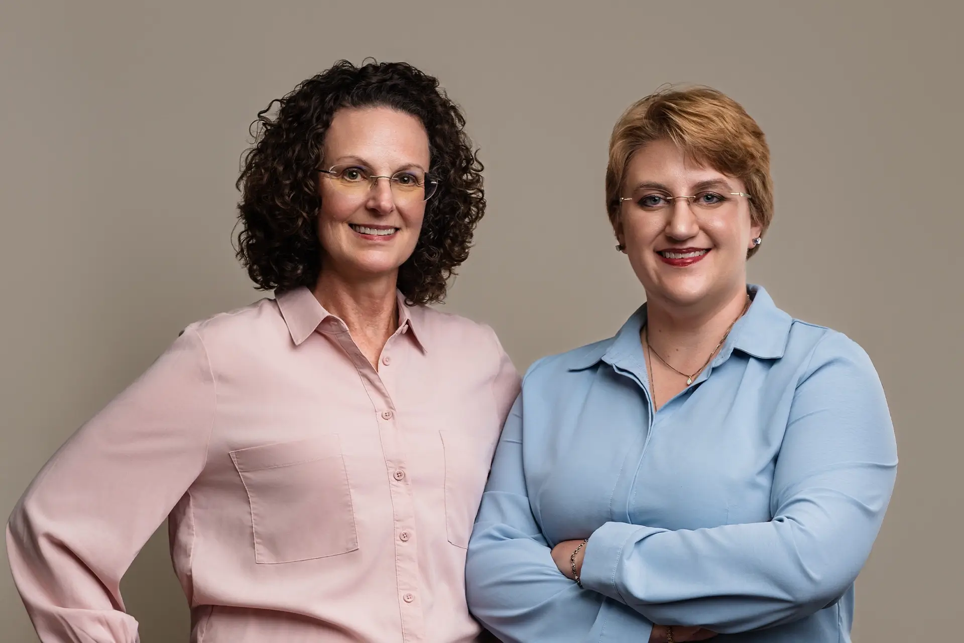 Two women, both smiling, stand side by side against a plain background. Perfect partners for small business bookkeeping Stillwater OK, one wears a pink blouse with curly hair; the other, in blue with glasses, is ready to support your financial needs. -Skiba Bookkeeping