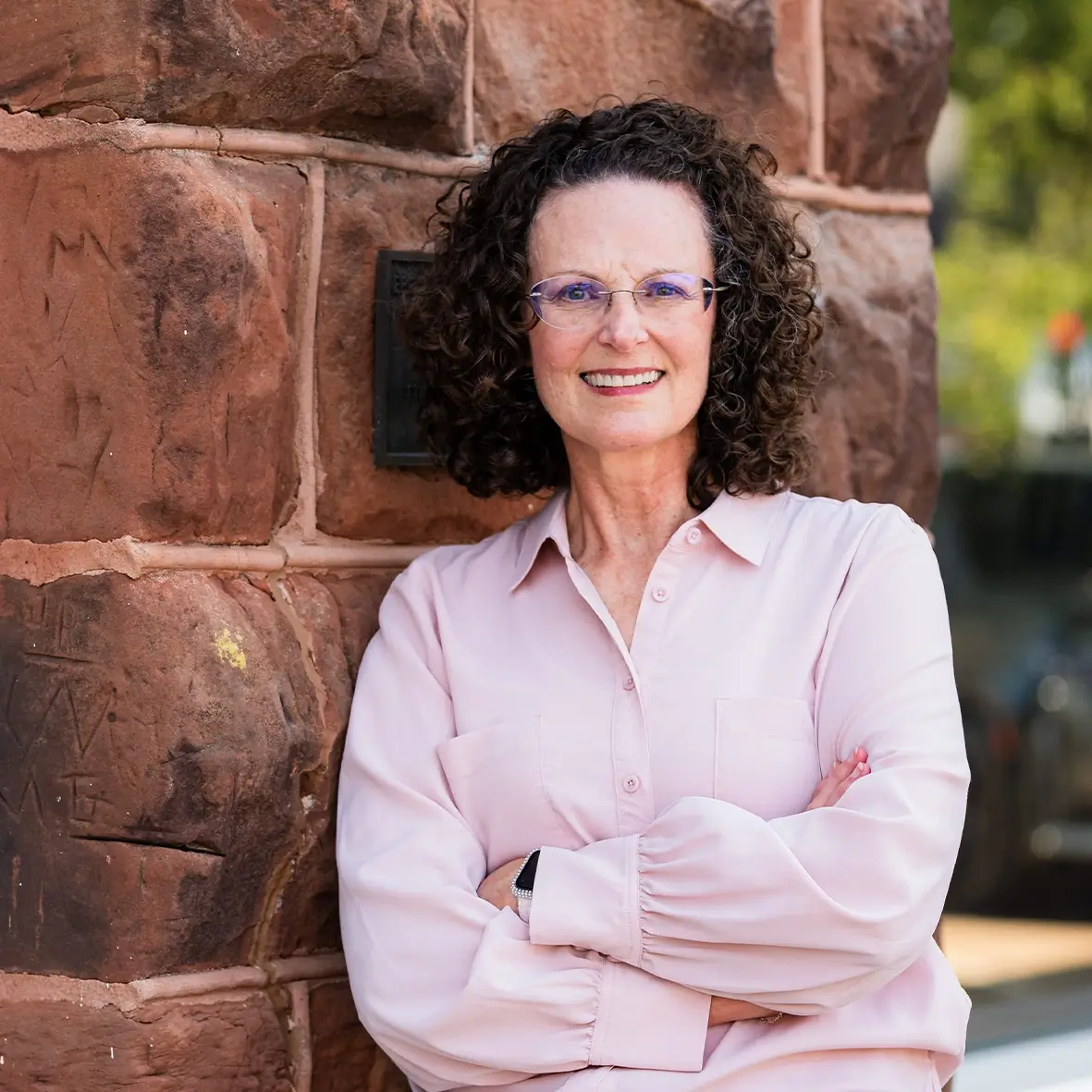 A woman with curly brown hair, glasses, and a light pink button-up shirt stands with arms crossed, leaning against a brown stone wall, smiling at the camera—your trusted bookkeeper for bookkeeping services in Stillwater OK. -Skiba Bookkeeping