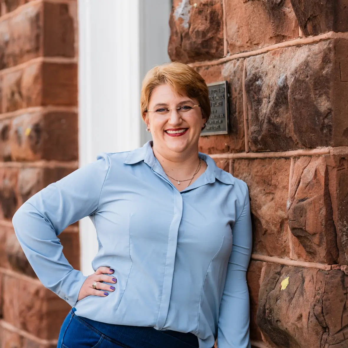 A person with short, light brown hair and glasses leans against a stone wall, smiling. They wear a light blue button-up shirt and dark pants—a friendly face ready to help with small business bookkeeping in Stillwater, OK. -Skiba Bookkeeping