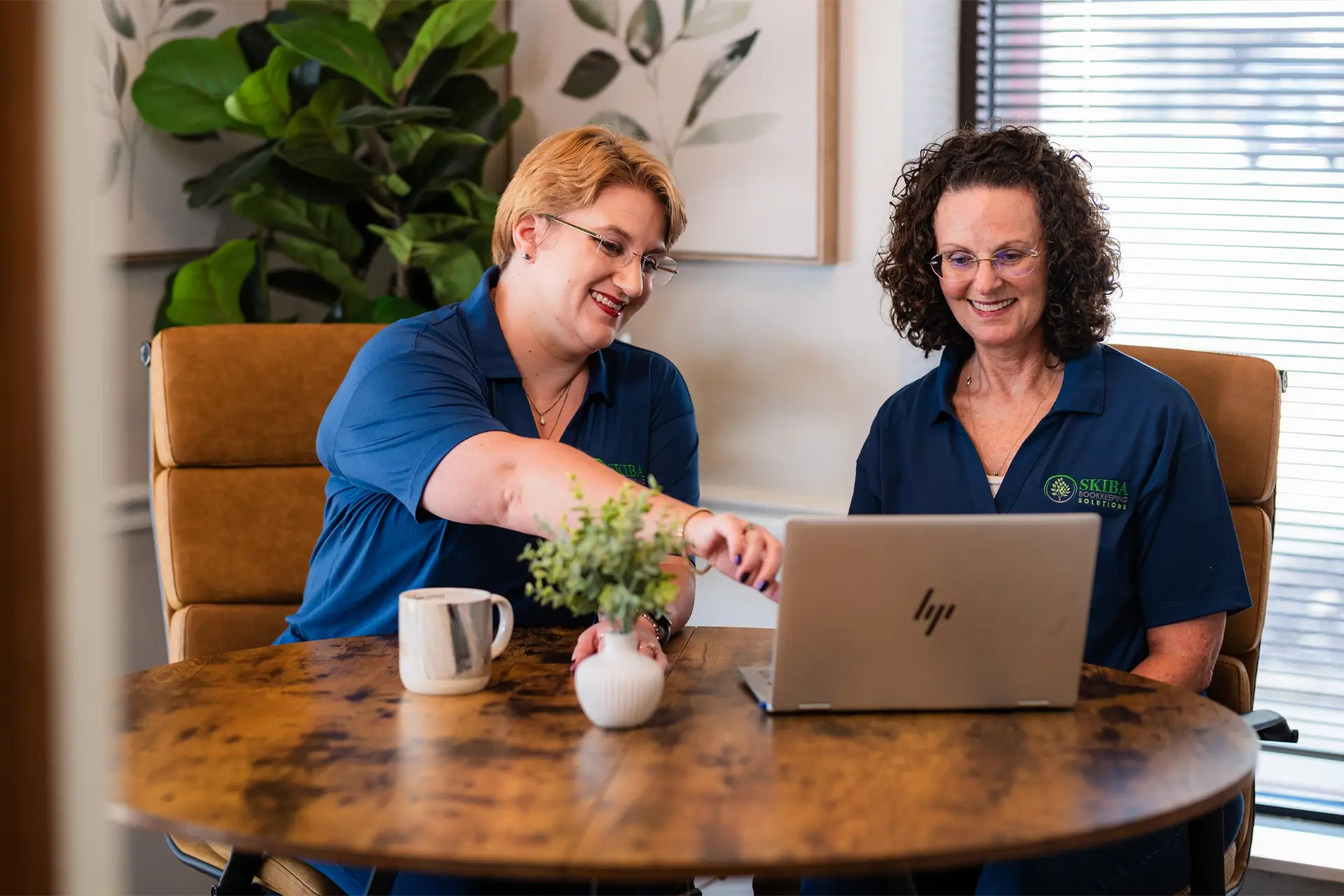 Two women sit at a round wooden table in Stillwater, OK, discussing small business bookkeeping as they look at a laptop. One points at the screen while the other smiles. A mug and small potted plant sit on the table, with a large plant in the background. -Skiba Bookkeeping