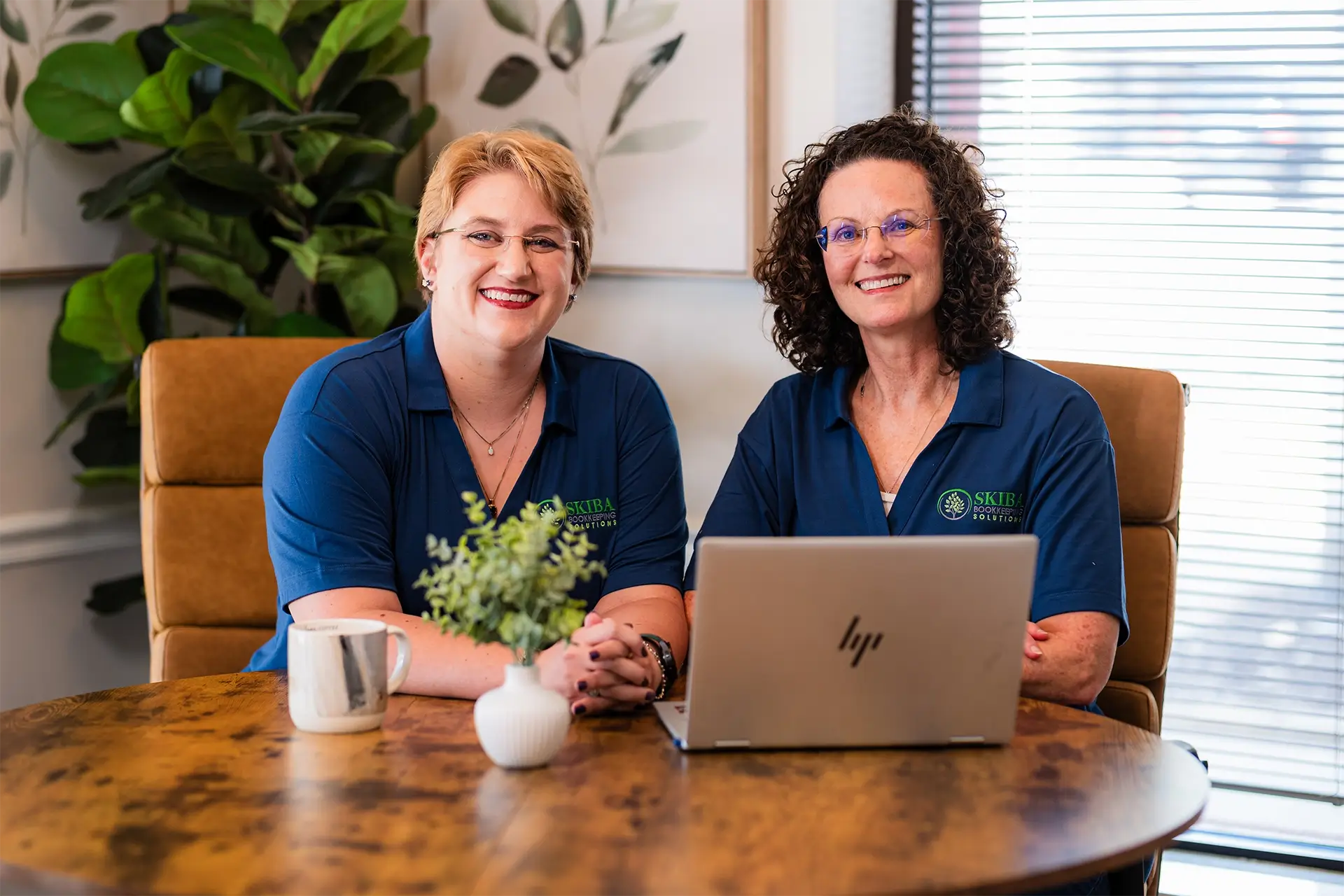 Two women wearing matching blue shirts sit at a round wooden table with a laptop, mug, and small plant—an inviting scene for bookkeepers offering small business bookkeeping in Stillwater OK. A large leafy plant and window blinds are in the background. -Skiba Bookkeeping